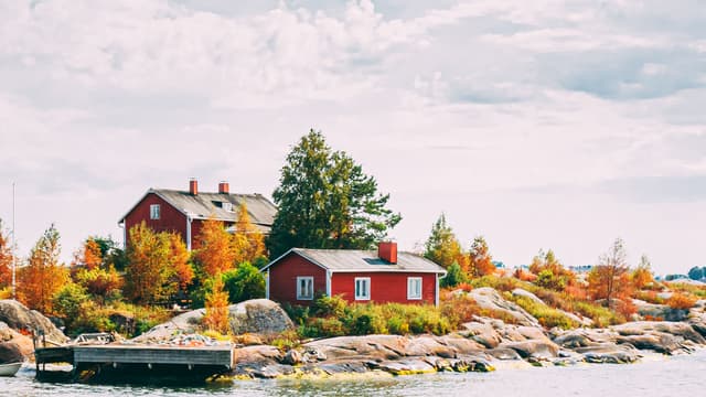 Kaksi punaista mökkiä ja laituri kauniissa saaressa jossa ruskan värittämiä puita.