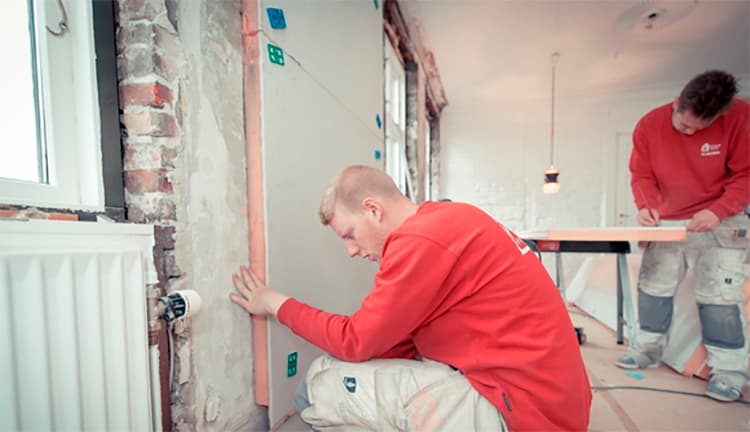 Oranssipaitainen mies asentaa huoneessa vanhan tiiliseinän päälle lisäeristettä
