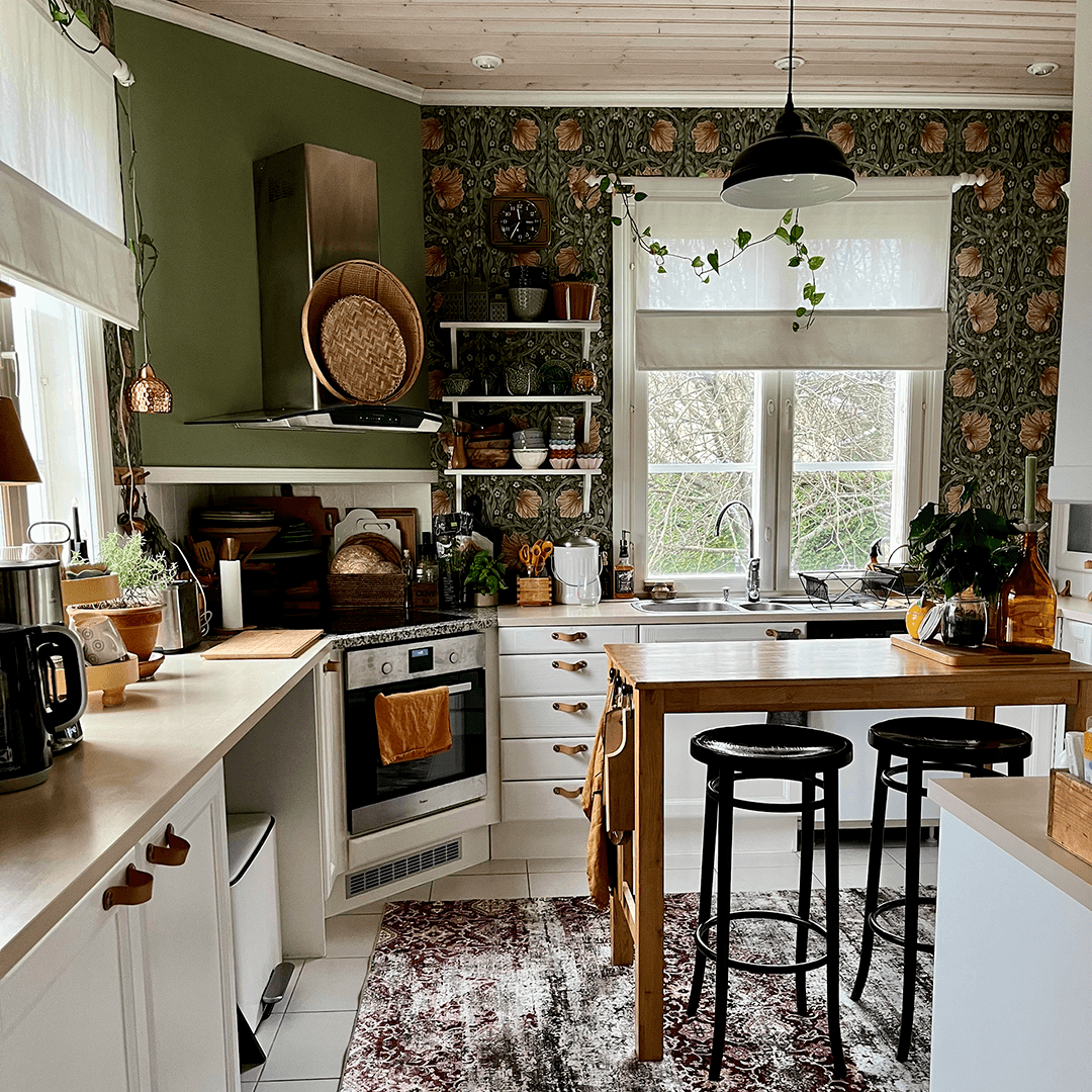 Kodikas ja runsassävyinen keittiö, jossa vihreät seinät, William Morrisin Pimpernel-tapetti, valkoiset kaapistot ja puinen saareke kahdella mustalla baarijakkaralla. Avohyllyillä on astioita ja koriste-esineitä, ja suuret ikkunat tuovat runsaasti luonnonv