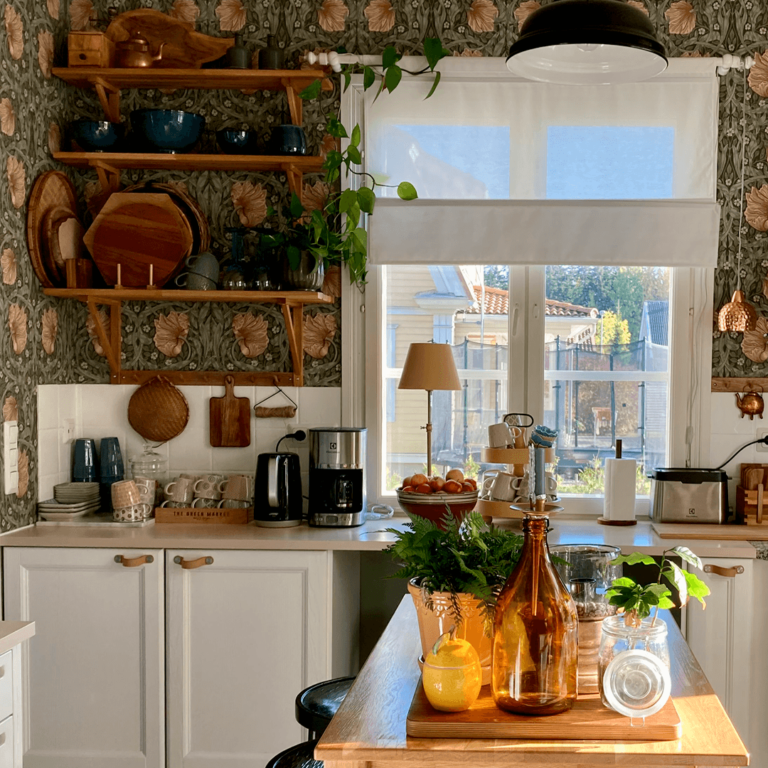 Keittiön nurkkaus, jossa Pimpernel-tapetti, puiset avohyllyt täynnä astioita ja kasveja, valkoiset kaapistot sekä suuri ikkuna, jonka eteen on asetettu hedelmävati ja koriste-esineitä. Luonnonvalo tulvii sisään ja valaisee puisen saarekkeen.