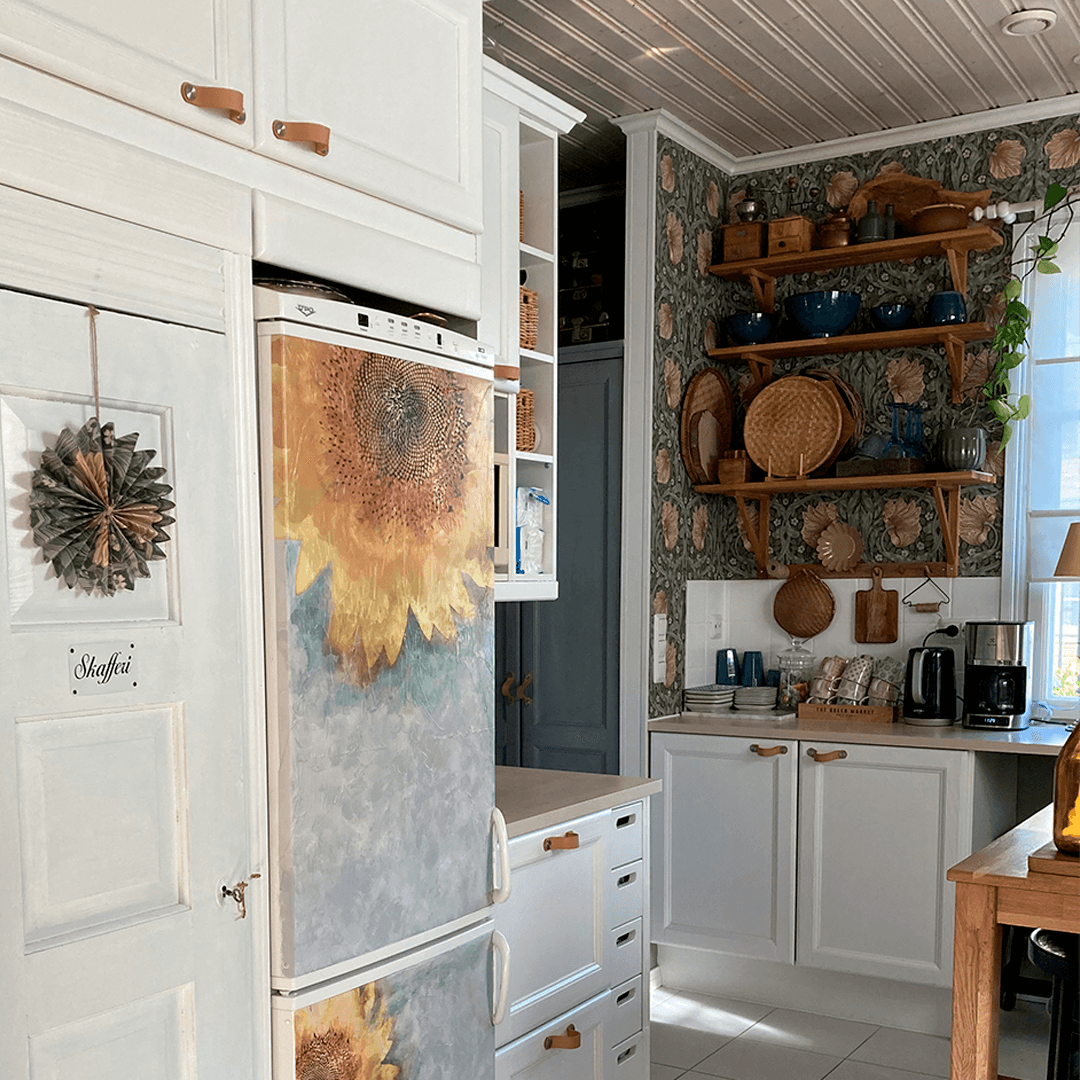 Keittiön nurkkaus, jossa valkoiset kaapistot, auringonkukalla koristeltu jääkaapin ovi ja puuhyllyt William Morrisin Pimpernel-tapettia vasten. Hyllyillä on astioita ja koreja, ja skafferin ovi näkyy vasemmalla.