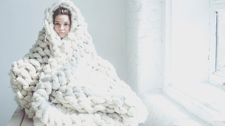 Nainen valkoista taustaa vasten paksuun kudottuun peittoon kääriytyneenä