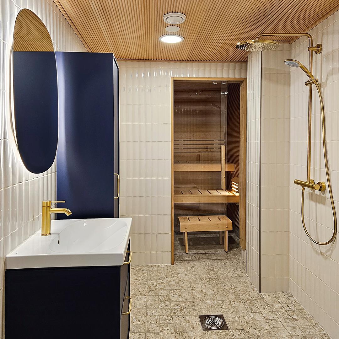 kuvassa nata salmelan remonttikohteen sauna ja pesuhuone. pesuhuoneessa on siniset kaapit, messinkinen suihku ja lasinen saunan liukuovi