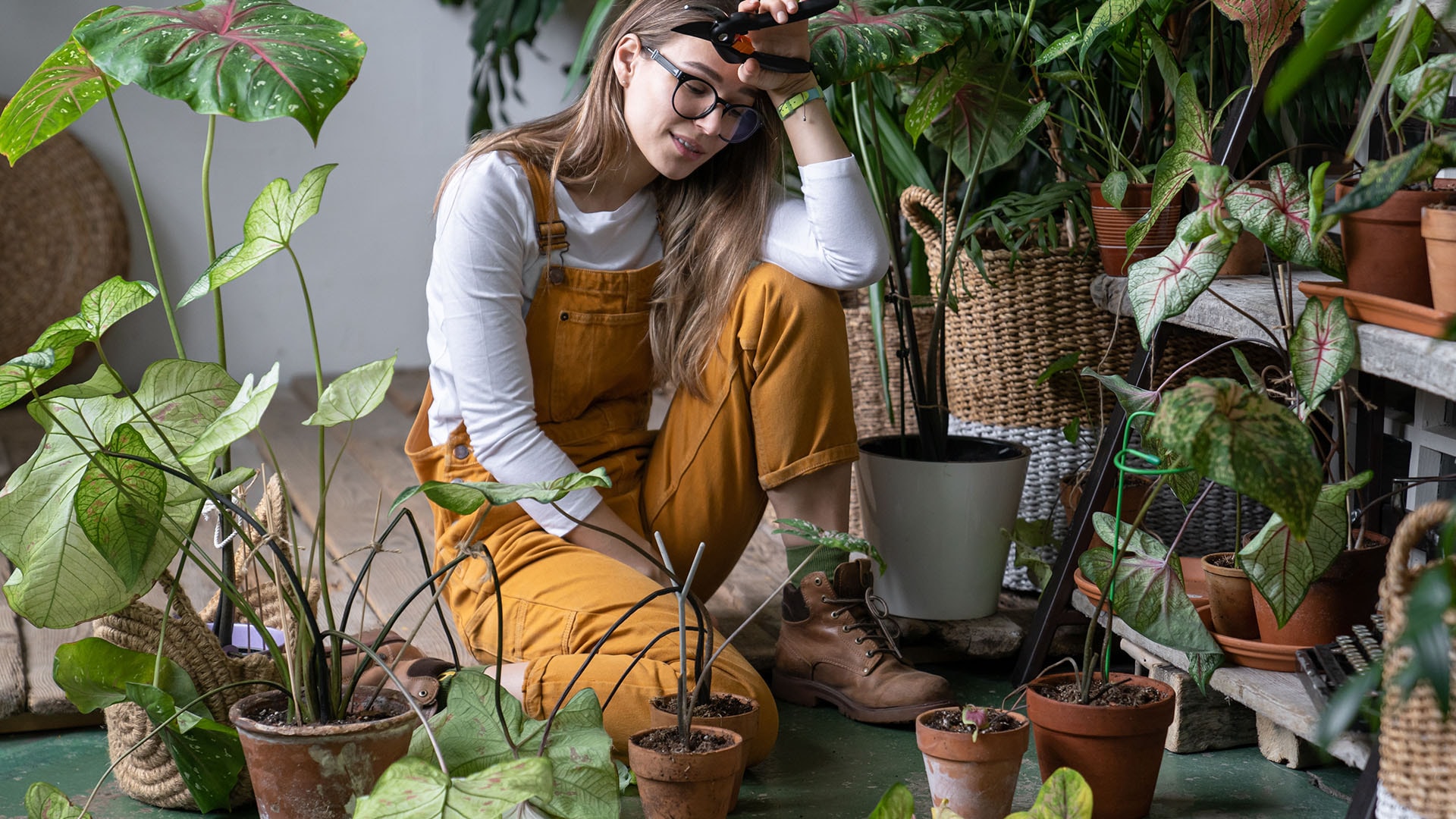 1920 nuori nainen istuu keltaisissa haalareissaan kukkien keskellä kädessään puutarhasakset