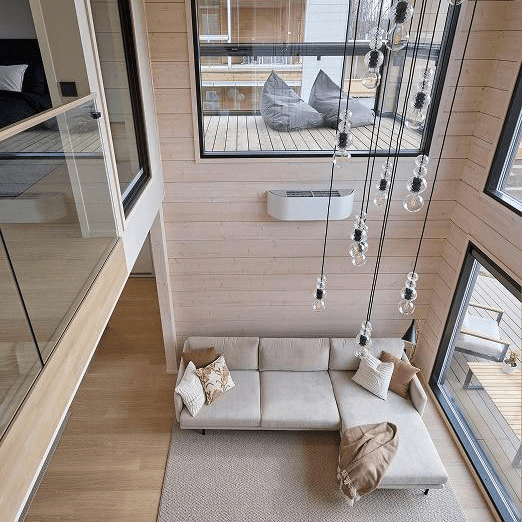 Kastellin hirsi Pesäpuu rakennuksesta kuva on sisältä yläkerrasta. Kuvassa näkyy suuret ikkunat mustilla karmeilla ja pääväri seinissä on vaaleanruskea. Kuvassa näkyy kattovalaistus joka on toteutettu yksittäisillä valopolttimoilla roikumaan ilmasta.