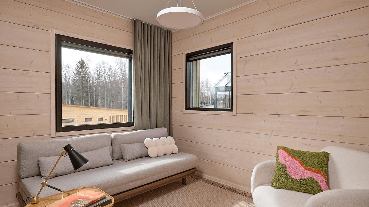 Kuvan seinien väri on vaalean hirren ruskea ja ikkunoiden mustat karmit pompsahtavat kokonaisuudesta ja sohvan ja pöydän sijoittelu luo tilan eloisuutta.