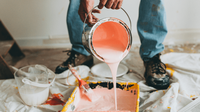 mies kaataa maalia maalikaukaloon jossa on maalaustela ja lattia on suojattu