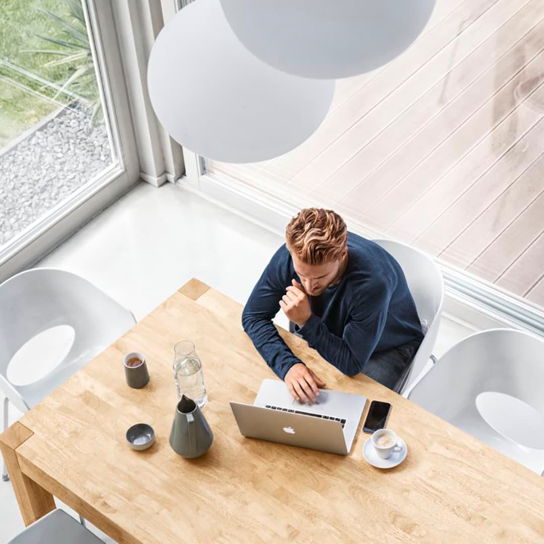 mies istuu pöydänvieressä läppärin kassa. kuvattu ylhäältäpäin ja suurista ikkunoista näkyy maisema