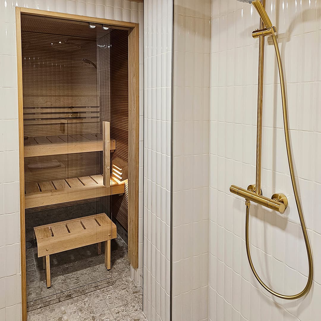 kirkaslasinen saunan liukuovi, valkoiset laatat pesuhuoneessa ja leppä panelointi sekä lauteet saunassa
