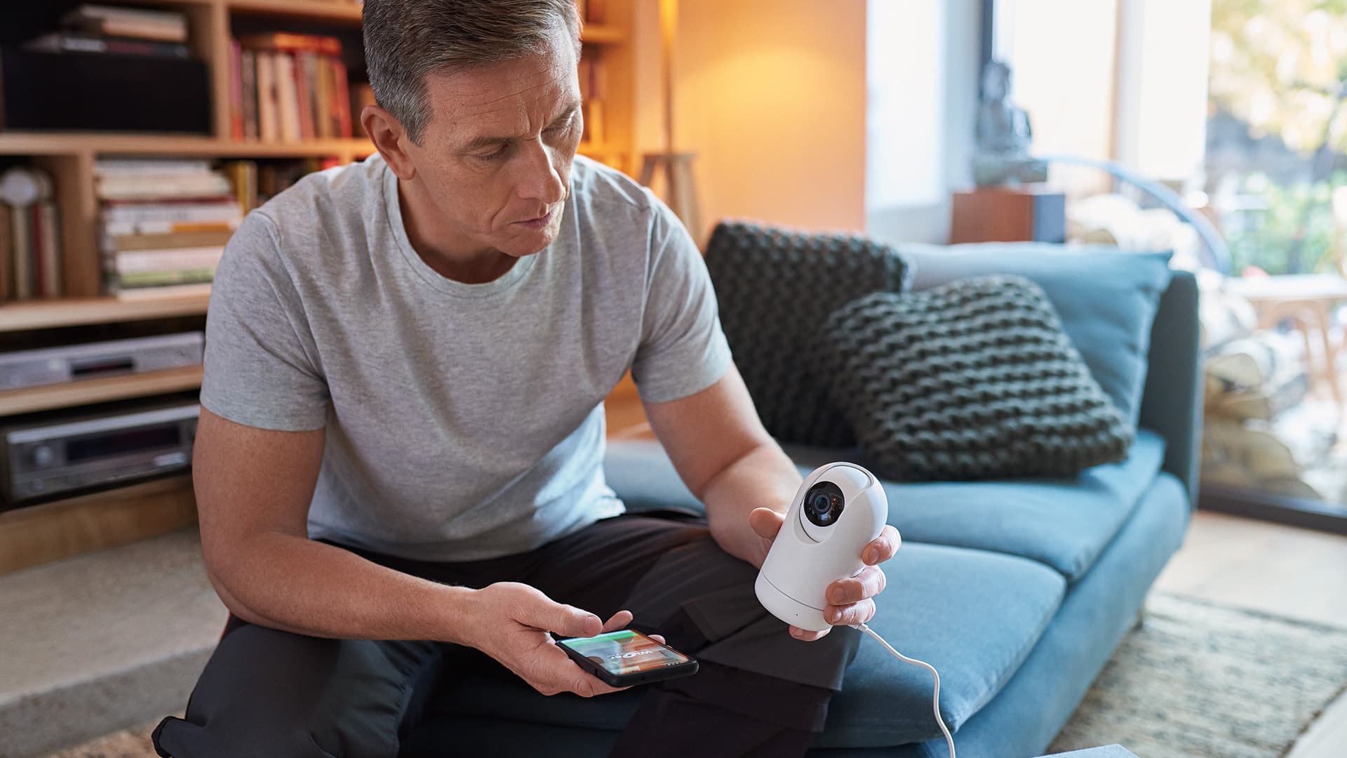 mies istuu olohuoneessa ja pitää kädessään valvontakameraa sekä älypuhelinta